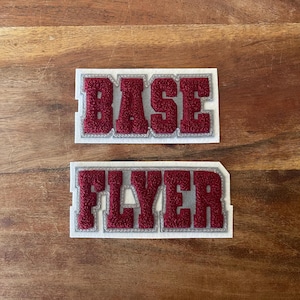 May include: Two maroon felt patches with white borders. The top patch reads "BASE" and the bottom patch reads "FLYER" in a varsity letter style. The patches are on a wooden surface.