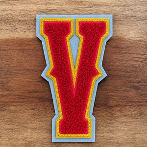 May include: A red chenille letter "V" patch with a gold border and light blue backing. The letter is centered on a wooden surface. The patch is likely for a jacket or bag.