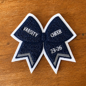 Może przedstawiać: Granatowa naszywka z szenili w kształcie kokardy cheerleaderskiej z białą filcową obwódką. Naszywka posiada biały haftowany napis VARSITY po lewej stronie oraz CHEER 23-26 po prawej stronie, z szarymi akcentami na dolnych końcach kokardy.
