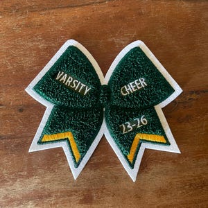 May include: A green and white cheerleading bow with the words "VARSITY CHEER" embroidered in gold thread. The bow also features the numbers "23-26" in gold. The bow is made of a textured material and has a white border.