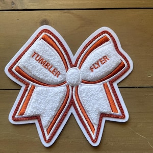 May include: A white and orange embroidered bow-shaped patch with the words "TUMBLER" and "FLYER". The patch has a textured white center and orange and white trim. The patch is on a wooden surface.