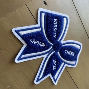 Puede incluir: Un parche bordado azul y blanco de lazo de animadora universitaria. El parche presenta las palabras "VARSITY", "CAPTAIN" y "CHEER", junto con el año "21-22". El lazo está sobre una superficie de madera.