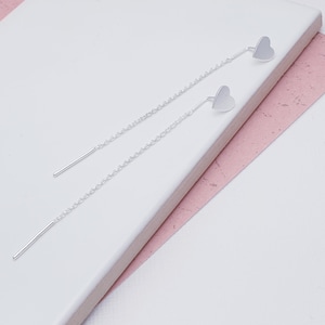 May include: A pair of silver heart-shaped dangle earrings with a delicate chain. The earrings are on a white surface with a pink surface behind it.
