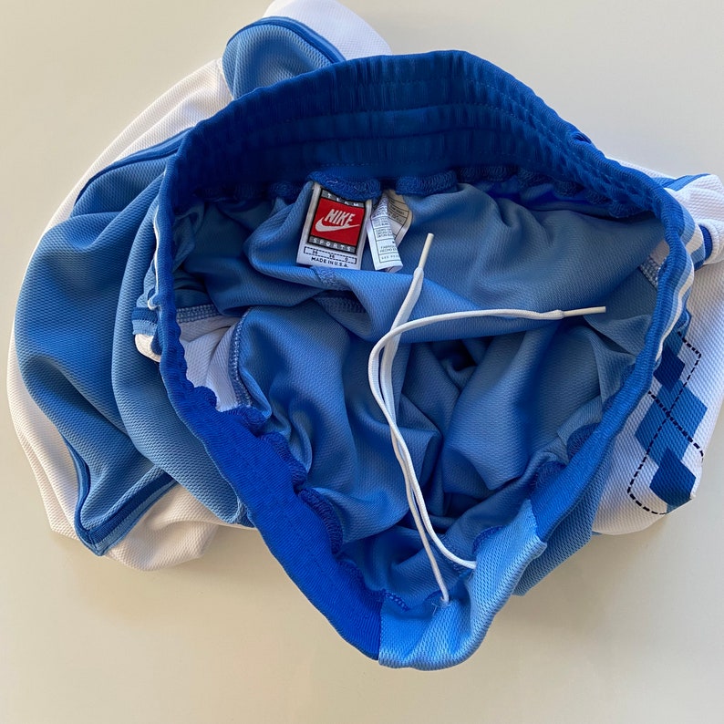 Op de afbeelding: Blauwe en witte sport shorts met een wit trekkoord en een Nike logo label. De shorts hebben een blauwe tailleband.