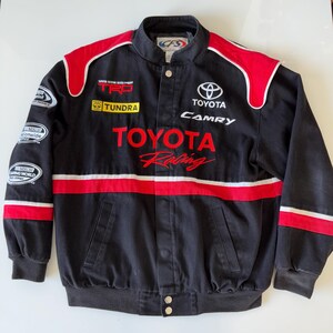 May include: Black Toyota Racing jacket with red and white stripes. The jacket features the Toyota logo, Camry, Tundra, and Toyota Racing logos.