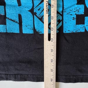 May include: Black t-shirt with large, turquoise text. A wooden ruler is positioned vertically along the right side, showing measurements in inches. The shirt's fabric is visible, with a focus on the text and the ruler's markings.