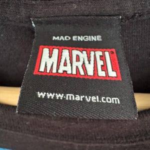 May include: Close-up of a black clothing tag with the text "MAD ENGINE" above a red and white "MARVEL" logo. Below the logo is the website address "www.marvel.com".
