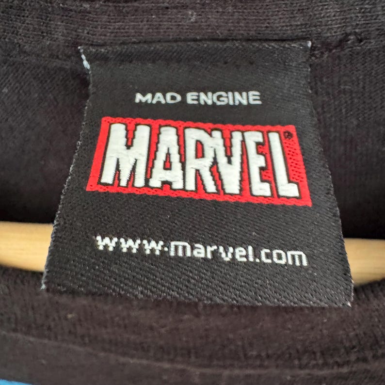 May include: Close-up of a black clothing tag with the text "MAD ENGINE" above a red and white "MARVEL" logo. Below the logo is the website address "www.marvel.com".