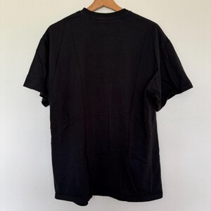 May include: A solid black t-shirt, viewed from the back, hanging on a wooden hanger. The shirt is made of a soft material and has short sleeves. The background is a plain white wall.