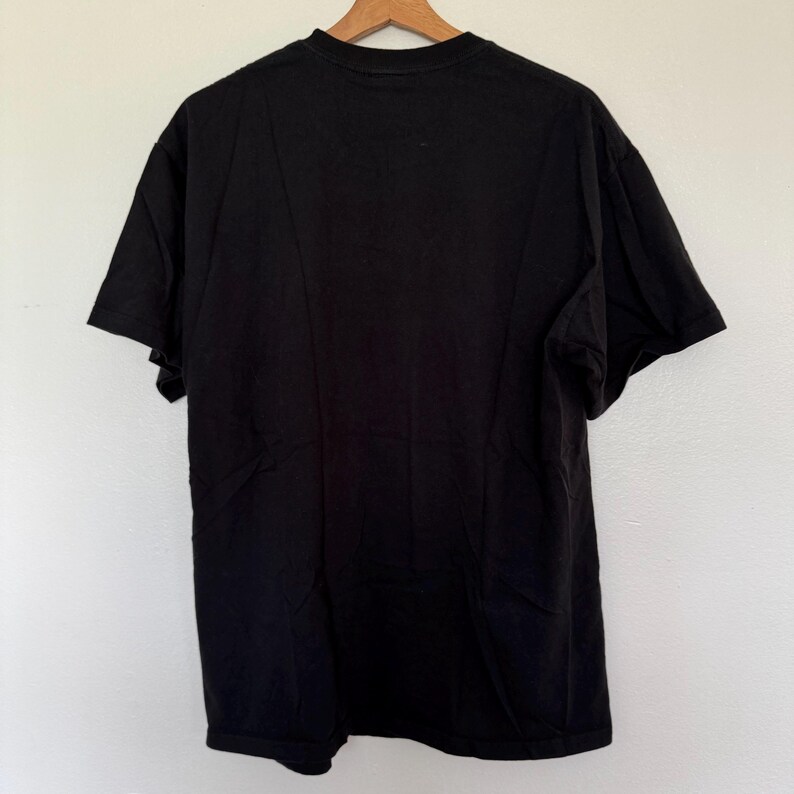 May include: A solid black t-shirt, viewed from the back, hanging on a wooden hanger. The shirt is made of a soft material and has short sleeves. The background is a plain white wall.