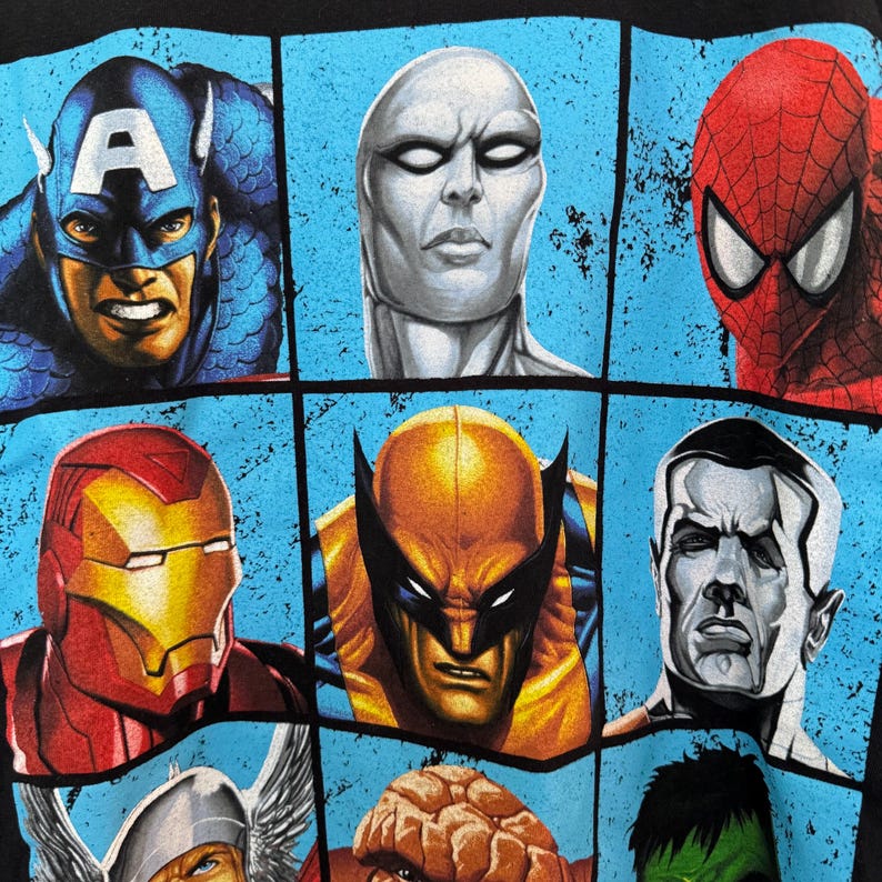 May include: Black t-shirt featuring a grid of comic book superhero faces. The grid includes Captain America, Silver Surfer, Spider-Man, Iron Man, Wolverine, and others. Each character is depicted in vibrant colors against a light blue background.