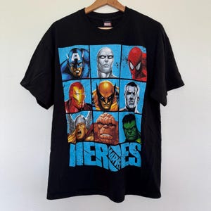 May include: Black t-shirt featuring a grid of Marvel superhero portraits, including Captain America, Spider-Man, and Hulk. The word "HEROES" is printed in blue below the portraits. The shirt is hanging on a wooden hanger.