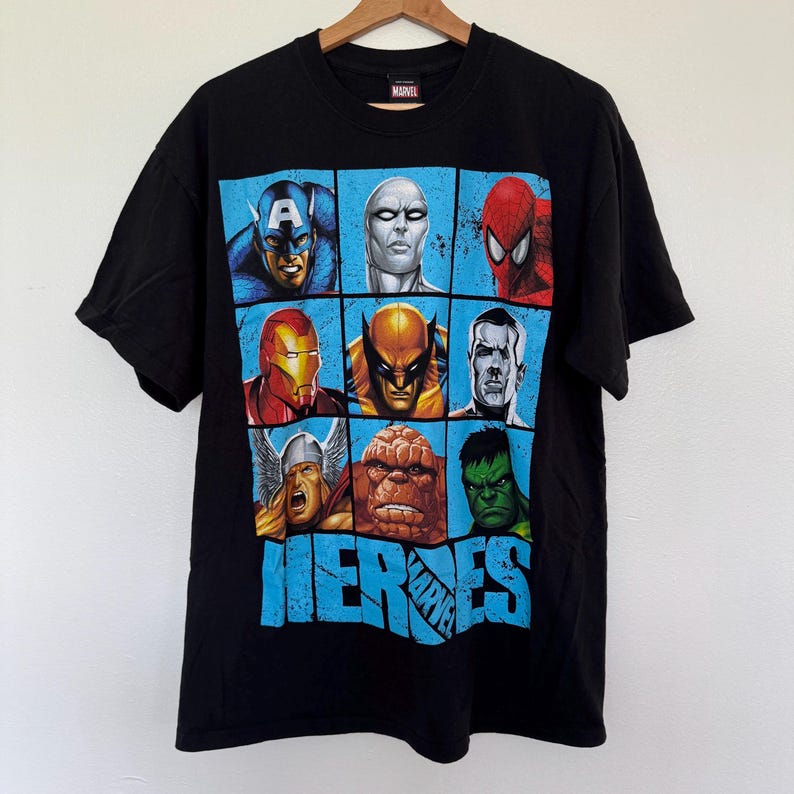 May include: Black t-shirt featuring a grid of Marvel superhero portraits, including Captain America, Spider-Man, and Hulk. The word "HEROES" is printed in blue below the portraits. The shirt is hanging on a wooden hanger.