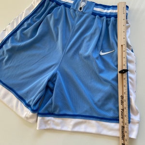 Op de afbeelding: Lichtblauwe basketbalshorts met witte bies en een Nike-logo. De shorts hebben een wit-blauw streepdessin op de tailleband en een klein wit voetafdruklogo aan de voorkant.