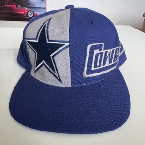 May include: A blue and gray baseball cap featuring the Dallas Cowboys logo. The cap has a split design with a star on the gray side and the word "Cowboys" on the blue side. The cap has a flat brim.