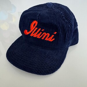 May include: A navy blue corduroy baseball cap with the word "Juini" embroidered in orange script across the front. The cap has a curved brim and a structured crown, suitable for casual wear.