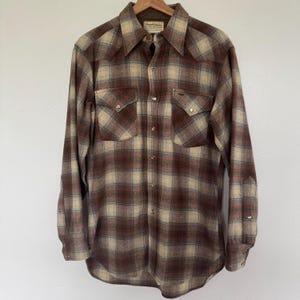 May include: A brown and beige plaid flannel shirt with a button-down collar and long sleeves. The shirt has two chest pockets with button closures and a label that reads "Penthouse" on the left side of the collar.