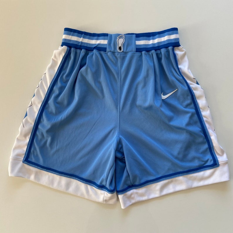 Op de afbeelding: Lichtblauwe basketbalshorts met witte en blauwe strepen op de tailleband, een witte Nike swoosh op het rechterbeen en een wit ruitpatroon aan de zijkanten.