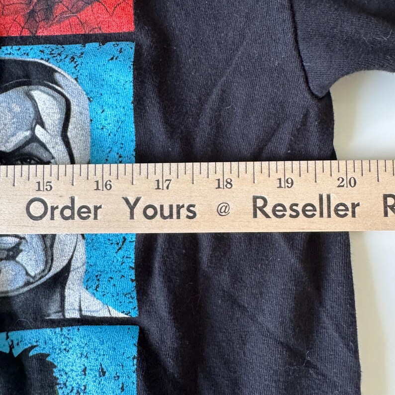 May include: Black t-shirt with a graphic design featuring a superhero character in red, blue, and gray. The text "Order Yours @ Reseller" is printed on a wooden ruler.