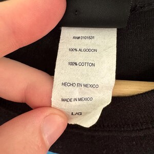 May include: A white tag with the text "RN# 0101531", "100% ALGODON", "100% COTTON", "HECHO EN MEXICO", "MADE IN MEXICO", and "L/G". The tag is attached to a black garment.