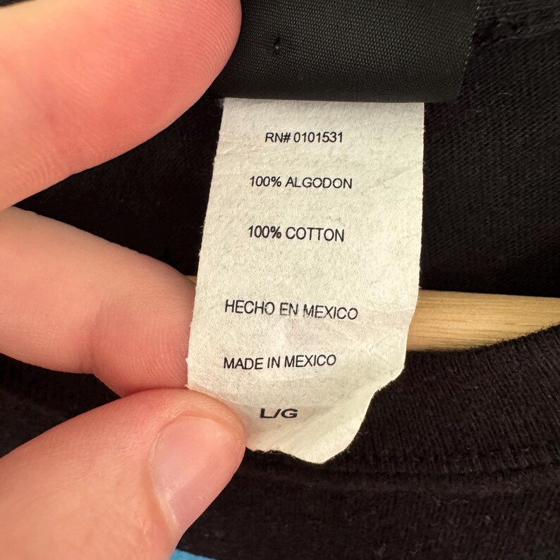 May include: A white tag with the text "RN# 0101531", "100% ALGODON", "100% COTTON", "HECHO EN MEXICO", "MADE IN MEXICO", and "L/G". The tag is attached to a black garment.