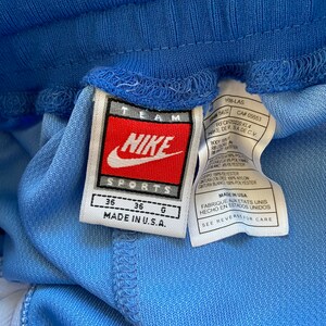 Op de afbeelding: Een wit label met rode en zwarte tekst met de tekst "TEAM NIKE SPORTS" en "MADE IN U.S.A." Het label is bevestigd aan een lichtblauwe sportbroek. Het label bevat ook maatgegevens: 36, 36, G. Het label bevat extra tekst in het Spaans en Frans.