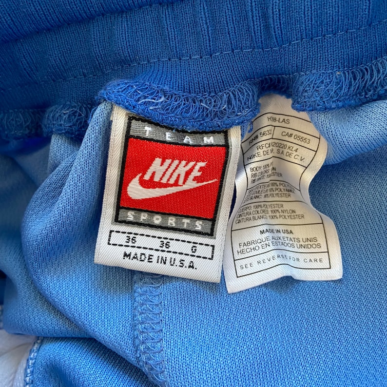 Op de afbeelding: Een wit label met rode en zwarte tekst met de tekst "TEAM NIKE SPORTS" en "MADE IN U.S.A." Het label is bevestigd aan een lichtblauwe sportbroek. Het label bevat ook maatgegevens: 36, 36, G. Het label bevat extra tekst in het Spaans en Frans.