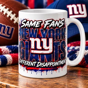 May include: A white ceramic mug featuring a graphic design with the text SAME FANS NEW YORK GIANTS DIFFERENT DISAPPOINTMENT and the NY logo in red, white, and blue. The design includes a paint splatter effect in blue and red colors.