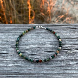 May include: A green, brown, and red beaded necklace with a silver clasp. The necklace is made of natural stones and is on a wooden surface.