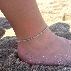 May include: Ankle bracelet made of small, light-colored beads. The beaded bracelet is worn on an ankle resting on sand. The beads are a mix of pale green, white, and tan. The background is a sandy beach.