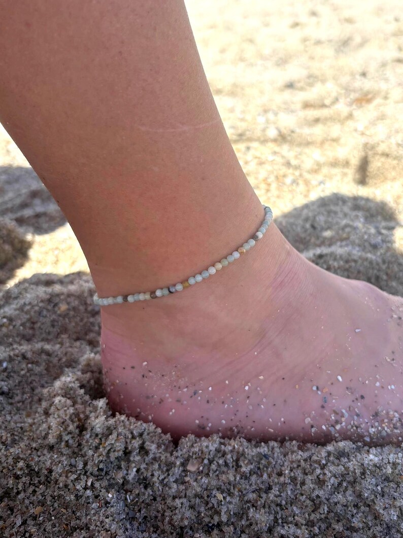 May include: Ankle bracelet made of small, light-colored beads. The beaded bracelet is worn on an ankle resting on sand. The beads are a mix of pale green, white, and tan. The background is a sandy beach.