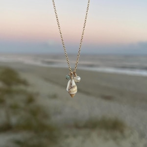 May include: A gold chain necklace featuring a seashell pendant and small pearl and green gemstone accents. The necklace is photographed against a blurred background of a beach.