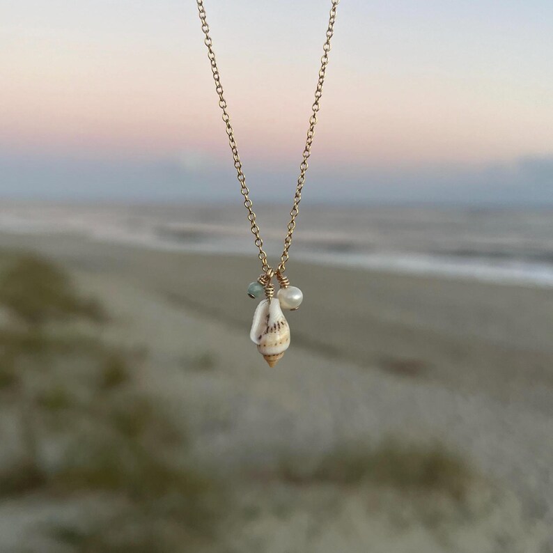 May include: A gold chain necklace featuring a seashell pendant and small pearl and green gemstone accents. The necklace is photographed against a blurred background of a beach.