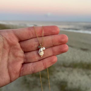 May include: A gold chain necklace featuring a seashell pendant with a small pearl and green gemstone accents. The necklace is shown on a hand against a beach backdrop.