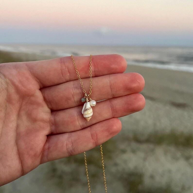 May include: A gold chain necklace featuring a seashell pendant with a small pearl and green gemstone accents. The necklace is shown on a hand against a beach backdrop.