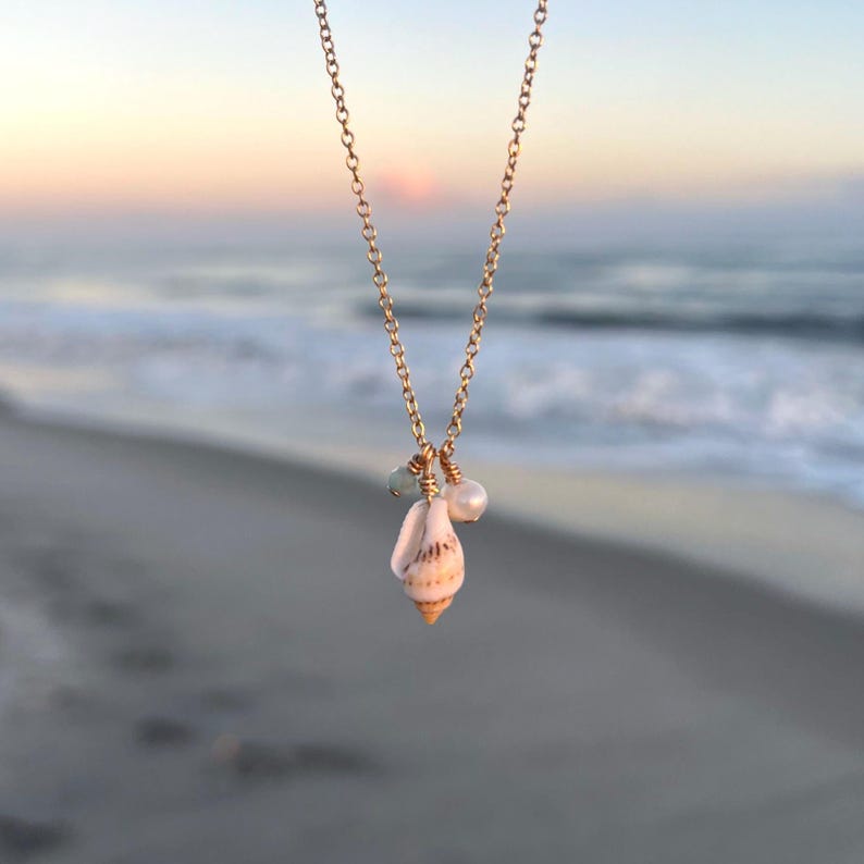 May include: A gold chain necklace with a seashell pendant and two small beads. The seashell is white with brown stripes and the beads are light blue and white.