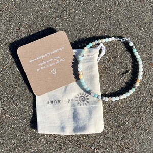May include: A light blue, green, and white beaded bracelet with a silver clasp. The bracelet is in a small white drawstring bag with the text "sunwraps" printed on it. A small card with the text "www.etsy.com/sunwraps made with love. on the coast of NC." is next to the bag.