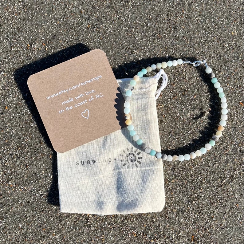 May include: A light blue, green, and white beaded bracelet with a silver clasp. The bracelet is in a small white drawstring bag with the text "sunwraps" printed on it. A small card with the text "www.etsy.com/sunwraps made with love. on the coast of NC." is next to the bag.