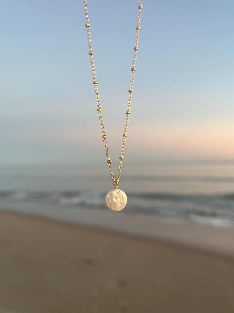 May include: A delicate gold chain necklace with a single, large, white pearl pendant. The necklace is photographed against a blurred background of a sandy beach and ocean.