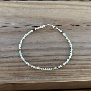 May include: A delicate beaded bracelet featuring small, round, light blue, brown, and black beads. The bracelet is strung on a thin wire and secured with a silver-colored clasp. The bracelet is displayed on a wooden surface.
