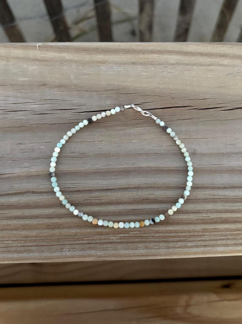 May include: A delicate beaded bracelet featuring small, round, light blue, brown, and black beads. The bracelet is strung on a thin wire and secured with a silver-colored clasp. The bracelet is displayed on a wooden surface.