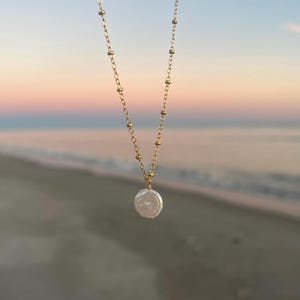 May include: A delicate gold chain necklace with a single, large, white pearl pendant. The necklace is photographed against a blurred background of a beach and sunset.