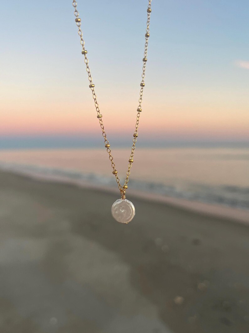 May include: A delicate gold chain necklace with a single, large, white pearl pendant. The necklace is photographed against a blurred background of a beach and sunset.