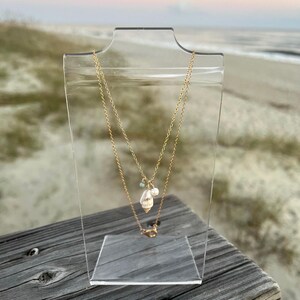 May include: A gold chain necklace featuring a seashell pendant and a pearl charm. The necklace is displayed on a clear acrylic stand against a beach backdrop.