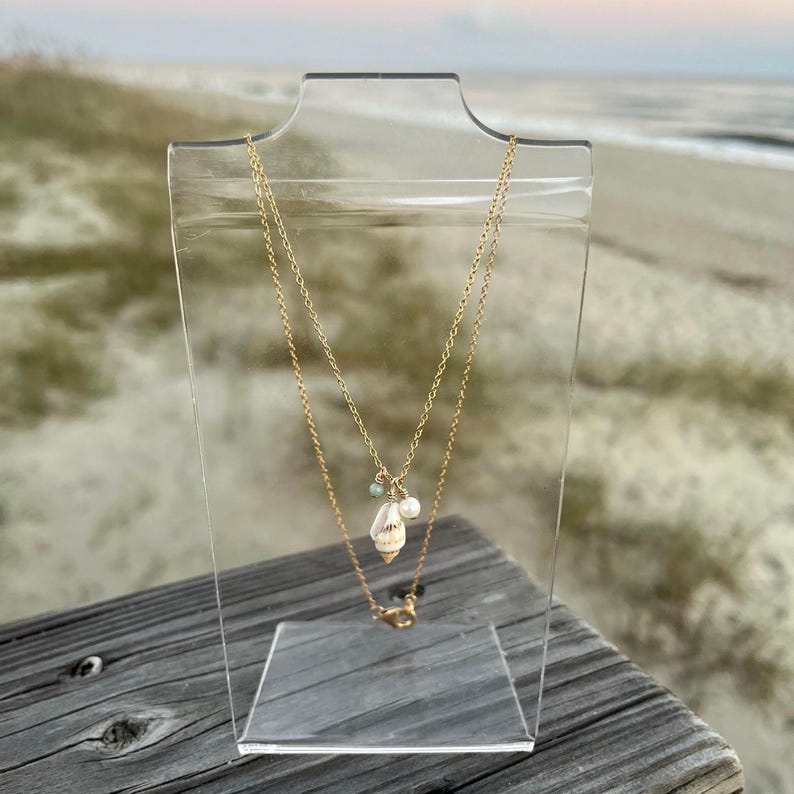 May include: A gold chain necklace featuring a seashell pendant and a pearl charm. The necklace is displayed on a clear acrylic stand against a beach backdrop.