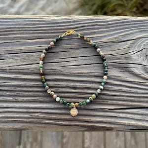 May include: A beaded bracelet featuring a gold sun charm. The bracelet is made of small, round, multi-colored beads in shades of green, brown, and pink. The bracelet is displayed on a weathered wooden surface.