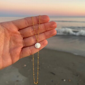 May include: A gold chain necklace with a white pearl pendant. The necklace is on a hand against a beach background.