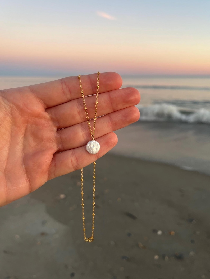 May include: A gold chain necklace with a white pearl pendant. The necklace is on a hand against a beach background.