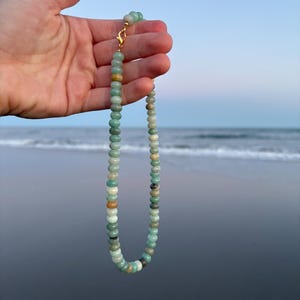 May include: A beaded necklace with a gold clasp, featuring a variety of round, colorful beads in shades of green, brown, and white. The necklace is held up against a backdrop of the ocean and sky.