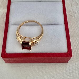 Op de afbeelding: Een gouden ring met een rechthoekige, dieprode edelsteen. De ring is gezet in een gouden band met kleine accentstenen. De ring wordt gepresenteerd in een rode sieradendoos met een witte binnenkant.